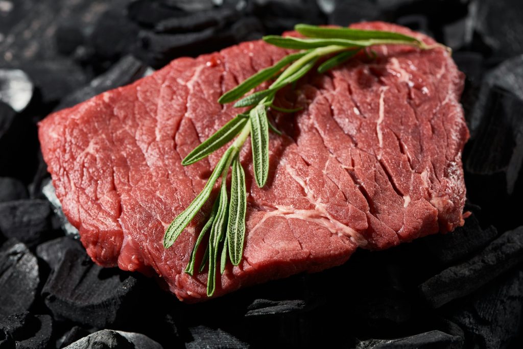 close up view of raw beef steak with rosemary twig on charcoals