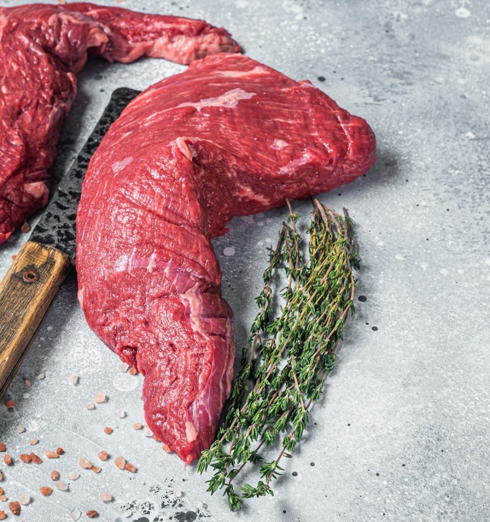 Unccoked tri-tip triangle roast, raw bottom sirloin steak on buther table with meat cleaver.